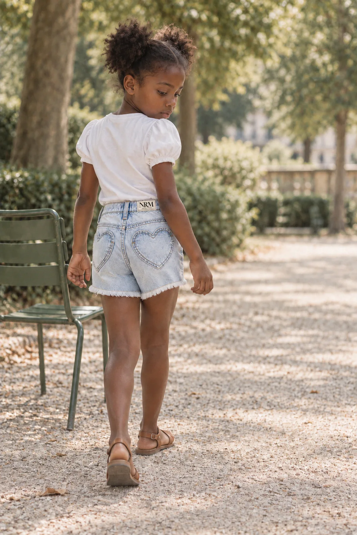 A girl in a white puff-sleeved tee and light heart-pocket denim shorts walks down a gravel path in a Paris park.