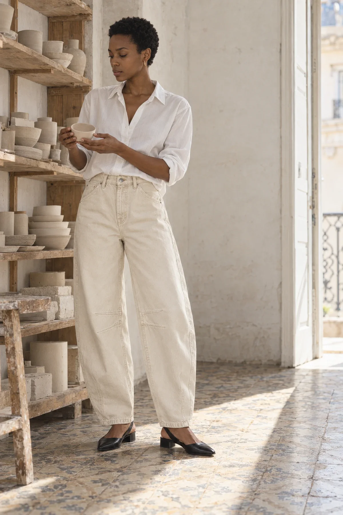 A woman in a white poplin shirt and ecru barrel jeans turns a small clay bowl in her hands in a ceramics studio in Lisbon.
