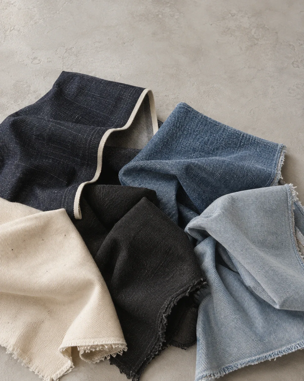 Cut lengths of denim draped over each other on pale plaster: raw indigo selvedge, mid-blue, washed light blue, black and ecru.